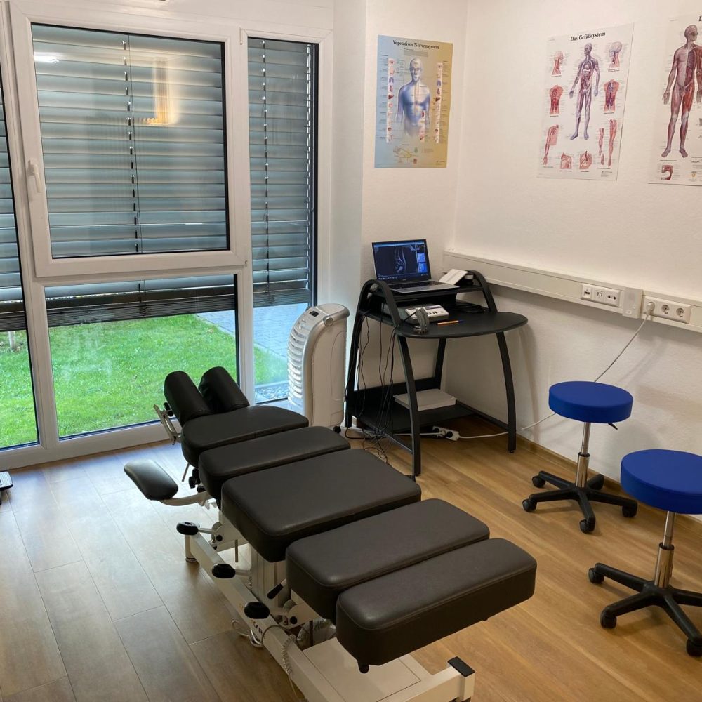 Blick in den Behandlungsraum der Chiropraxis Ulm mit professioneller Justierliege, anatomischen Postern des Nervensystems und der Station für den Wirbelsäulenscan.